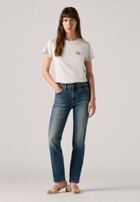 Grijze T-shirt met korte mouwen en een klein logo, gecombineerd met lichtblauwe rechte jeans. Model draagt beige sandalen met blokhak en bandjes.