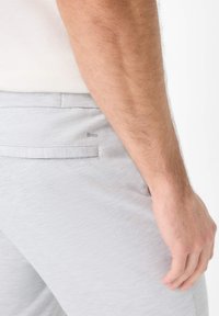Pantaloni grigi chiari da uomo in tessuto testurizzato, dotati di una tasca posteriore e un logo sottile vicino alla cucitura della tasca. Senza chiusure visibili.