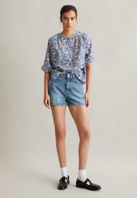 Bloemmotieven, blouse met korte mouwen in blauw en roze, gecombineerd met hoge denimshorts. Model draagt zwarte schoenen met witte sokken.