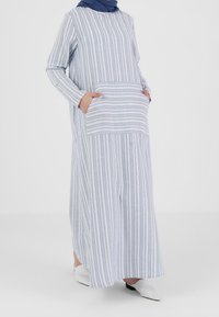 Robe longue rayée en bleu clair et blanc, avec des manches longues et une poche sur le devant. Fabriquée en tissu léger avec une texture lisse.