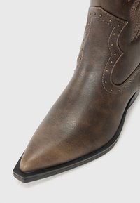 Bota de cowboy de cuero marrón con punta afilada, costuras detalladas y tachuelas de tono dorado en el costado, con una superficie texturizada y suela negra.