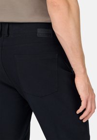Pantalones negros para hombre hechos de tejido elástico. Cuentan con un bolsillo parche y una etiqueta de logo de goma en la parte trasera. Textura suave y ajuste entallado.