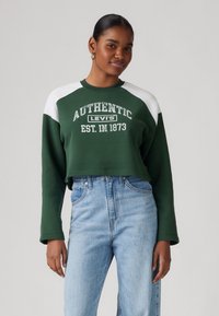 Zelená skrátená mikina s bielymi raglánovými rukávmi, na prednej strane s potlačou „AUTHENTIC LEVI'S EST. IN 1873“, v kombinácii so svetlomodrými džínsami.