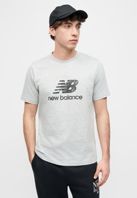 Νέος άνδρας με μαύρο καπέλο, που φοράει γκρι μπλουζάκι New Balance και μαύρο παντελόνι, στέκεται με τα χέρια στις τσέπες, κοιτάζοντας δεξιά.