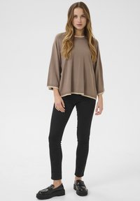Bruine oversized pullover met een ronde hals en beige afwerking, gecombineerd met zwarte skinny jeans en zwarte platformloafers. Glad textuur.