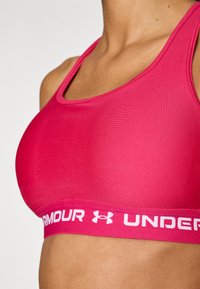 Hladká športová podprsenka v horúcej ružovej farbe so strihom racerback a kontrastným bielym logom Under Armour na texturovanej pružnej časti.