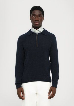 Jersey de punto azul marino con cuello con cremallera, puños acanalados y dobladillo. Lleva puesto un camisa de color verde claro con cuello, con un acabado texturizado.
