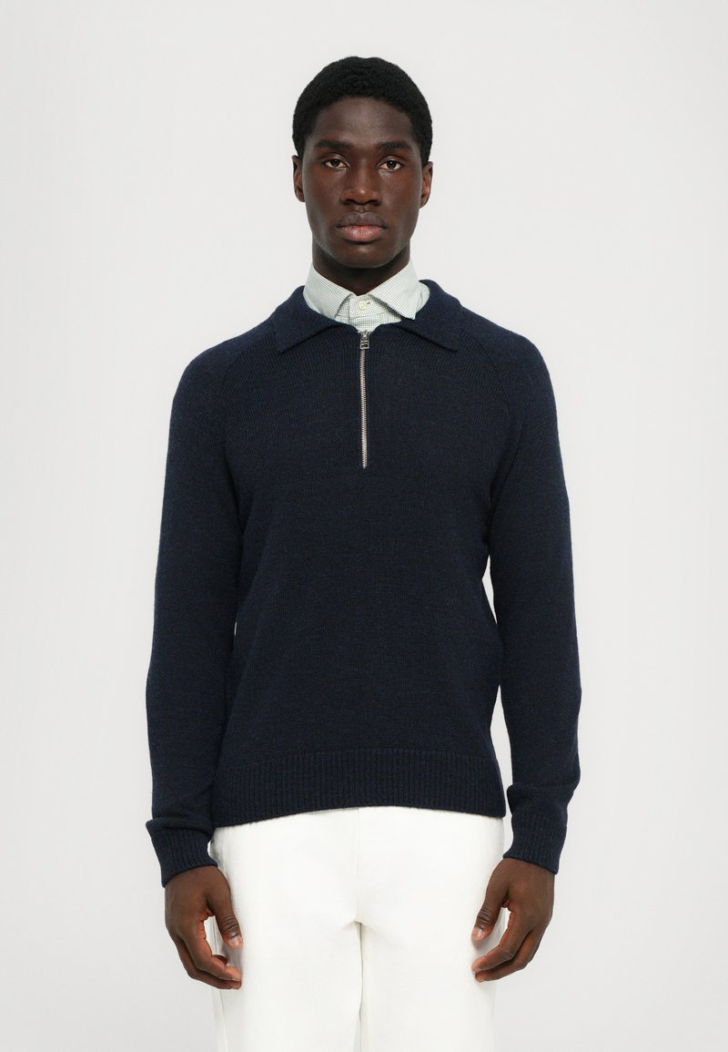 Jersey de punto azul marino con cuello con cremallera, puños acanalados y dobladillo. Lleva puesto un camisa de color verde claro con cuello, con un acabado texturizado.