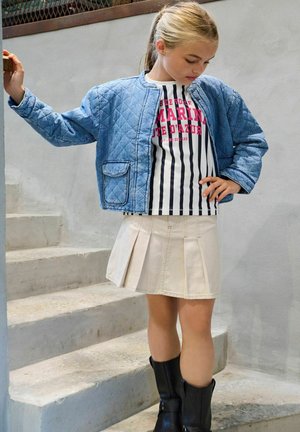 Noor tüdruk sinist tikk-kangast jope, triibulise graafilise t-särgi, beežil volditud seeliku ja mustade saabastega, seismas betoonist trepil.