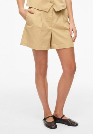 Žena oblečená v béžových šortkách na mieru a rovnakej vesti, stojaca s jednou rukou v vrecku, obutá v čiernych balerínach Mary Jane.
