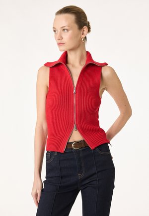 Femme portant un haut rouge côtelé sans manches avec fermeture éclair et col, jean taille haute foncé, et ceinture marron, debout avec une main sur la hanche.