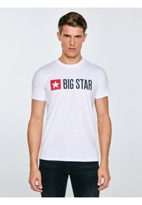 Fiatal férfi fehér pólóban, amelyen piros csillag logó és fekete "BIG STAR" felirat látható, fekete nadrággal, egyszínű háttér előtt állva.