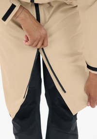 Beige waterdichte jas met zwarte ritssluiting, gedeeltelijk geopend. De textuur lijkt glad, met een contrasterende donkerder stof aan de onderrand en mouwen.