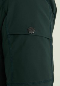 Temno zelene kratke hlače za prostočasne aktivnosti z teksturirano površino. Imajo velik stranski žep in podrobnost v obliki okroglega črnega kovinskega gumba.