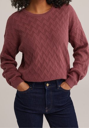 Femme portant un pull mauve texturé avec des motifs géométriques et un jean bleu foncé, les mains décontractées dans les poches.