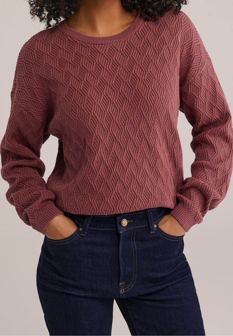 Femme portant un pull mauve texturé avec des motifs géométriques et un jean bleu foncé, les mains décontractées dans les poches.