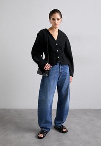 Černý strukturovaný blazer s dlouhým rukávem a hlubokým výstřihem do V, zapínání na přední knoflík, v kombinaci s volnými modrými džíny a sandály s otevřenou špičkou se cvočky.