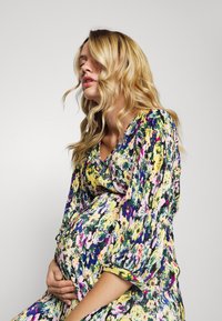 Vestido de maternidade floral com cores vivas, textura plissada e mangas longas bufantes, projetado para conforto e estilo durante a gravidez.