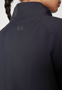 Detailní záběr na horní část zad osoby v černé bundě Under Armour s vyvýšeným límcem a pletenými vlasy.