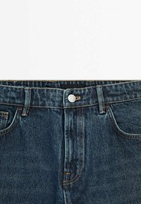 Dunkelblaue Jeans mit silbernem Knopf, Gürtelschlaufen, Vordertaschen und sichtbaren Nähten auf weißem Hintergrund.