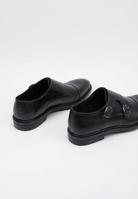 Zapatos de cuero negro con un acabado liso, que presentan dos hebillas metálicas circulares, punta redondeada y una suela de goma texturizada y baja.