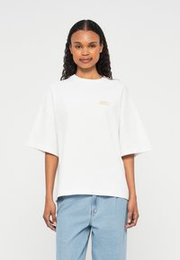 Λευκό oversized T-shirt από βαμβάκι, με στρογγυλή λαιμόκοψη και μικρό χρυσό λογότυπο στην αριστερή πλευρά. Το μοντέλο φοράει μπλε τζιν παντελόνι.