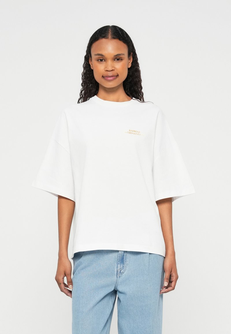 Λευκό oversized T-shirt από βαμβάκι, με στρογγυλή λαιμόκοψη και μικρό χρυσό λογότυπο στην αριστερή πλευρά. Το μοντέλο φοράει μπλε τζιν παντελόνι.