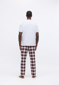 Άνδρας που στέκεται ξυπόλητος με την πλάτη προς τα πίσω, φορώντας ένα απλό λευκό κοντομάνικο πουκάμισο και κόκκινα-λευκά καρώ παντελόνια σε λευκό φόντο.