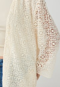 Spitzen-Open-Cardigan in Creme mit floralen und geometrischen Mustern. Verfügt über lange, weite Ärmel und ein leichtes, strukturiertes Design.