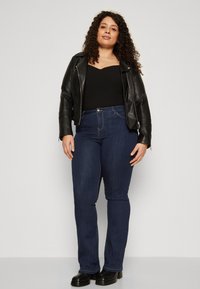 Chaqueta de cuero negra de moto sobre una camiseta negra ajustada, combinada con jeans azul oscuro y botas negras robustas. Cabello rizado enmarcando la cara.