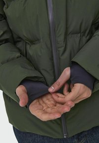 Chaqueta de plumas verde con cierre de cremallera, que presenta puños oscuros, aberturas elásticas en las muñecas y una textura acolchada. Manos posicionadas cerca de la chaqueta.