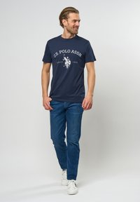 Navyblaue Baumwoll-T-Shirt mit weißem Text, versehen mit einem Logo und einer Grafik eines Polo-Spielers. Kombiniert mit blauen Jeans und weißen Turnschuhen.