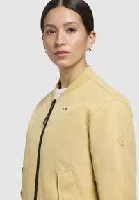 Kollane pommikindel jakk, mis on valmistatud siledast kangast, millel on ees lukuhaak, küljetaskud ja ümmargune logo varrukal.