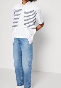 Kortärmad vit skjorta med marinblå horisontella ränder, tillsammans med ljusblå jeans och vita sneakers, mot en enfärgad bakgrund.