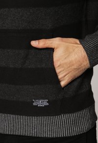 Czarny i szary sweter w paski z kieszenią z przodu, ściągaczami na mankietach i u dołu. Posiada metkę marki na pasie z instrukcjami prania.