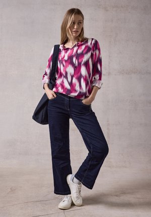 Multikleurige blouse in roze en wit met een V-hals, opgerolde mouwen; donkerblauwe wijde jeans; witte sneakers; zwarte totebag.