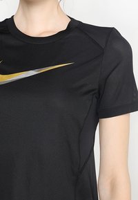 Musta urheilu T-paita, joka on valmistettu kevyestä ja hengittävästä kankaasta; siinä on verkkotekstuuria ja heijastava hopea- ja kultainen Nike swoosh -logo.