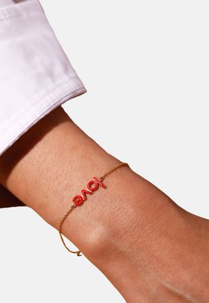 Bracelet en chaîne dorée avec un texte rouge épelant "amour", avec une surface lisse et polie sur un poignet contre un fond clair.