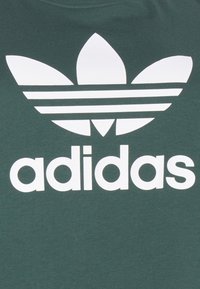 Grön bomullströja med en stor vit Adidas-logotyp med tre ränder och en bladdesign, främst centrerad på tyget.
