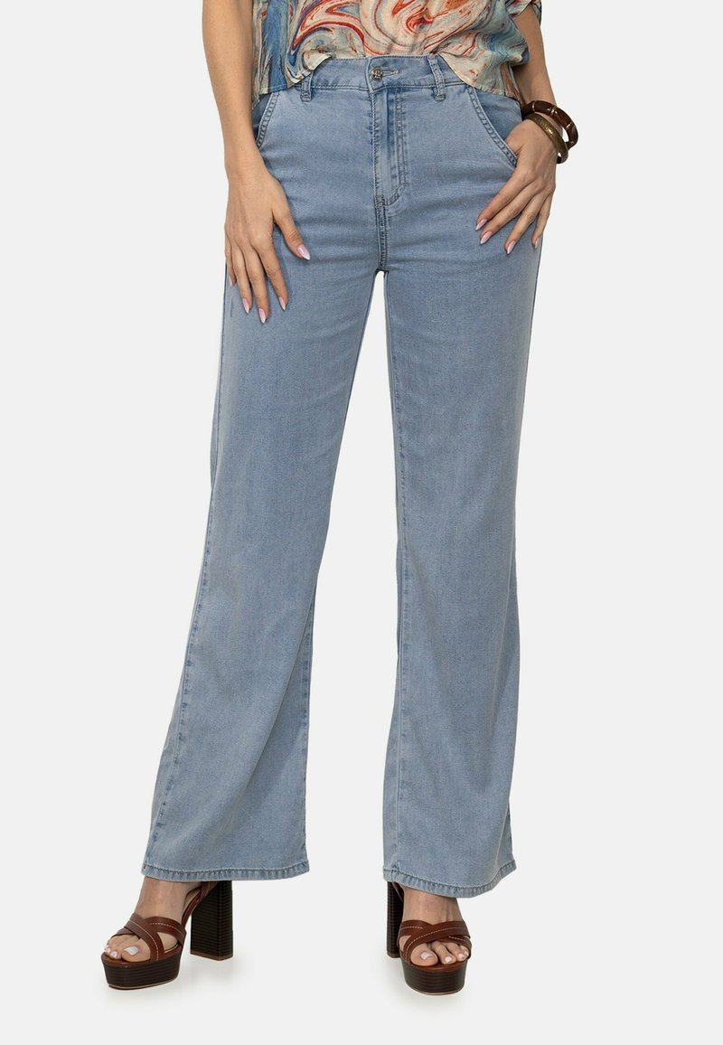Jeans de pierna ancha color azul claro con cintura alta, presentan bolsillos delanteros y cierre de botón. Combinados con sandalias de tacón marrones.