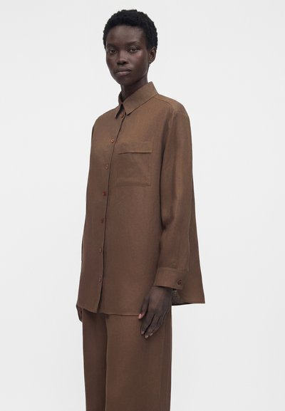 Γυναίκα με χαλαρό καφέ πουκάμισο με κουμπιά και τσέπη στο στήθος, καθώς και παντελόνια σε φαρδιά γραμμή, στέκεται μπροστά σε απλό φωτεινό φόντο.