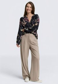 Blouse met lange mouwen in een donkere stof met bloemmotief, gecombineerd met lichtbeige broek met wijde pijpen en witte sneakers.