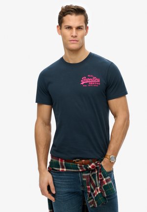 Navy blue cotton t-shirt with crew neck and short sleeves, featuring pink SuperDry logo on the upper left chest. Jeans and plaid shirt tied at the waist.