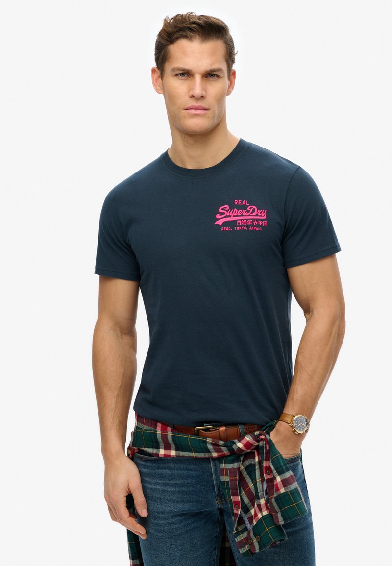 Camiseta de algodón azul marino con cuello redondo y mangas cortas, con el logo de SuperDry en rosa en el pecho superior izquierdo. Jeans y camisa de cuadros atada en la cintura.