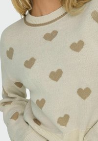 Strickpullover in Creme mit braunen Herzmustern, ausgestattet mit einem gerippten Halsausschnitt und strukturiertem Stoff. Das Design umfasst lange Ärmel und eine lockere Passform.