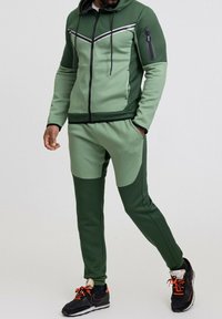 Ensemble de sport vert comprenant un hoodie zippé avec des rayures blanches contrastantes et un pantalon de jogging. Fabriqué en tissu lisse avec un design ajusté.