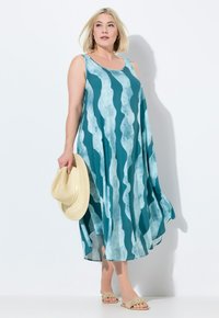 Ein ärmelloses, türkisfarbenes Kleid mit einem wellenförmigen Muster in helleren Farbtönen, aus leichtem Stoff, fließendem Design, kombiniert mit einem Strohhut und Sandalen.