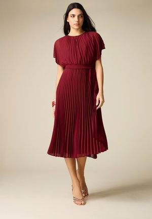 Nainen, joka pukeutuu hihattomaan, laskostettuun viininpunaiseen midi-mekkoon, jossa on vyötäröä korostava malli, seisoo yksinkertaisen beigen taustan edessä.