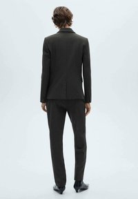 Tmavě šedý oblek na míru s texturovaným povrchem, vybavený jedním rozparkem vzadu a sladěnými zúženými kalhotami. Hladká, elegantní obuv.