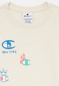 Camiseta de algodón beige con cuello redondo, con logotipos coloridos de Champion, texto "NEW YORK" y detalles de llamas en azul, rosa y verde.